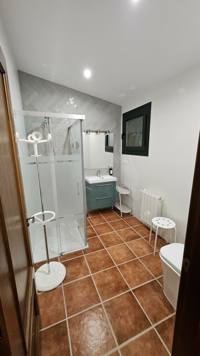 Baño moderno con ducha acristalada, lavabo verde, suelo terracota y ventana negra